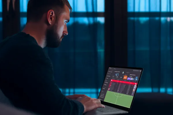 একজন পুরুষ তার ল্যাপটপে মনোযোগ দিয়ে কাজ করছেন, ব্যাকগ্রাউন্ডে লাইভ স্ট্যাটস — যা 2777BED মোবাইল অ্যাপের স্পোর্টস ও গেমিং ফিচারগুলোকে তুলে ধরে।